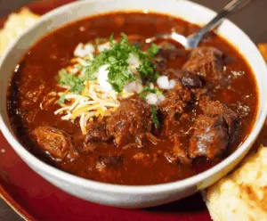 A steaming bowl of authentic Texas brisket chili topped with shredded cheese and chopped onions, served with cornbread on the side.
