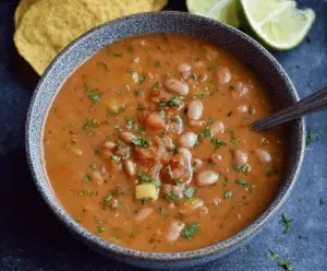A bowl of Mexican Pinto Bean Soup topped with fresh cilantro, diced tomatoes, and a squeeze of lime, served with warm tortilla chips on the side.
